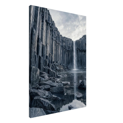 Waterfall cascading between tall basalt columns into a reflective pool, with dark foreground rocks and a cloudy sky.