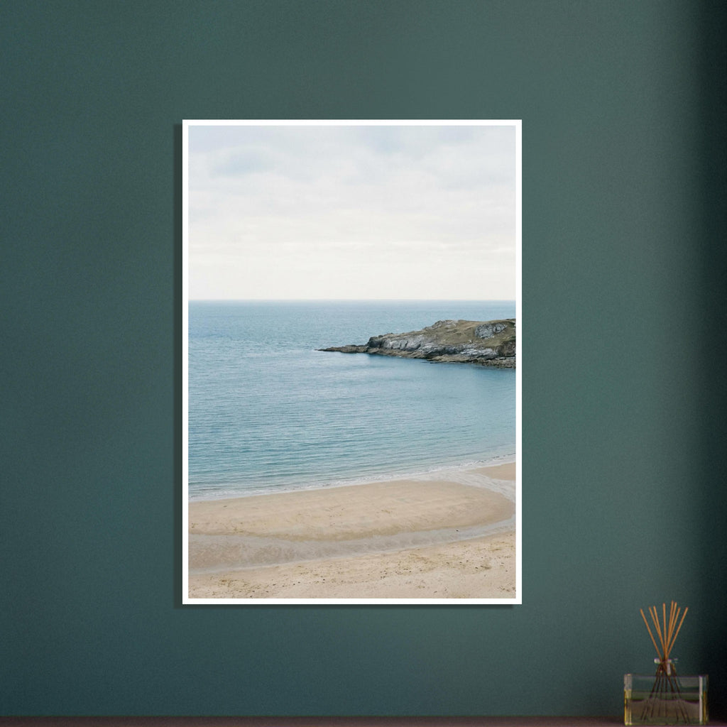 Sandy bay with calm blue water, a rocky headland and a curving tidal channel beneath a pale, overcast sky.