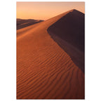 Desert sand dune with rippled orange slopes, a sweeping ridge and deep brown shadow beneath a peach sky.