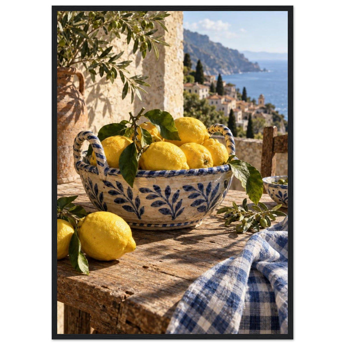 Lemons in a blue-and-white bowl on a wooden table, with olive branches, checked cloth and a coastal village beyond.