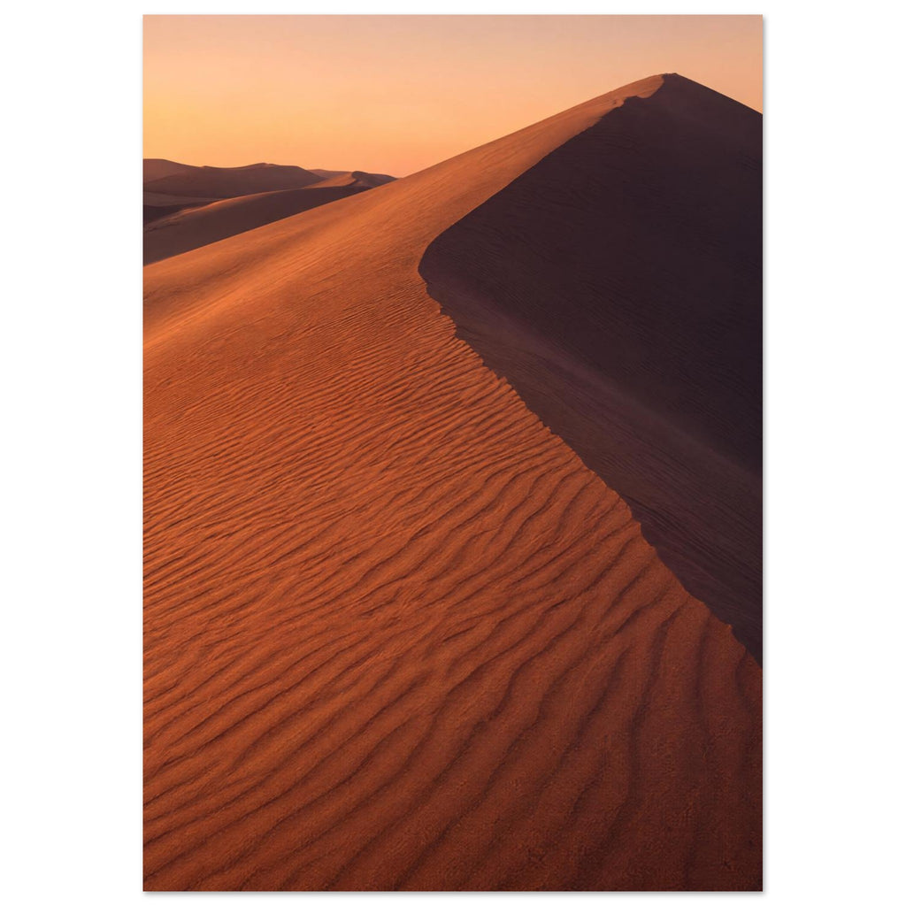 Desert sand dune with rippled orange slopes, a sweeping ridge and deep brown shadow beneath a peach sky.