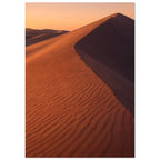 Desert sand dune with rippled orange slopes, a sweeping ridge and deep brown shadow beneath a peach sky.