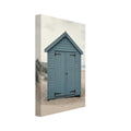 Blue wooden beach hut with closed doors on pale sand, with dune grasses, sea waves and an overcast sky.