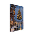 Illuminated Christmas tree suspended from a harbour crane above a snowy quay, with boats and golden reflections in blue water