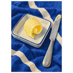 The Butter Curl still life showing a curled pat of butter on a rectangular dish beside a silver knife on cobalt cloth.
