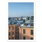 Snow-covered green rooftops and chimneys above peach buildings, with a distant city skyline beneath a clear blue sky.