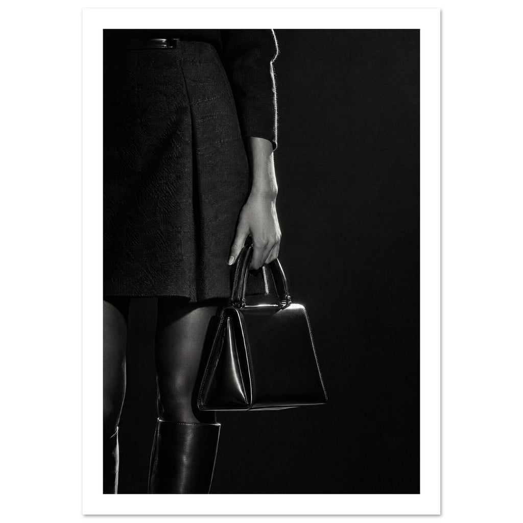 Cropped black and white photograph of a figure in a dark skirt and boots holding a glossy structured handbag.