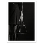 Cropped black and white photograph of a figure in a dark skirt and boots holding a glossy structured handbag.