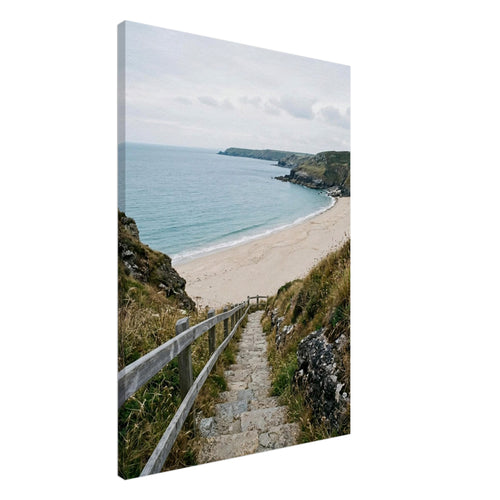Stone steps with a wooden handrail descend through coastal grasses to a sandy beach, blue sea and rocky headlands.