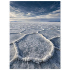 Salt flats with raised polygon-shaped ridges stretching towards distant mountains beneath a blue, cloud-streaked sky.