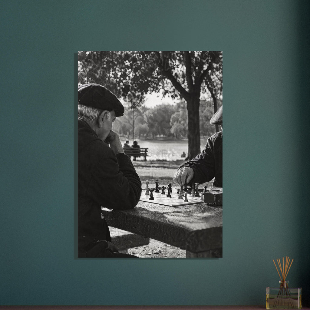 Two men in flat caps playing chess at a stone table beside water and trees, photographed in black and white.