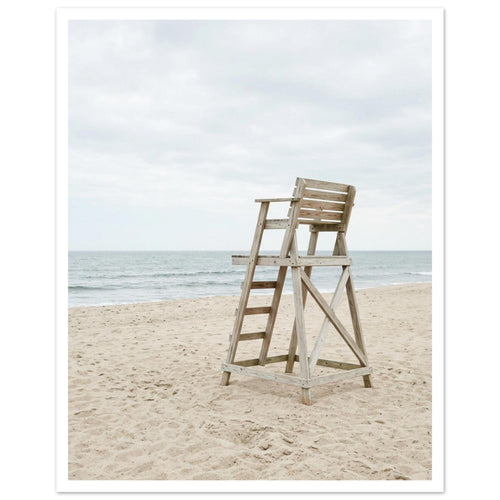 Empty wooden lifeguard chair on pale sand beside gentle sea waves beneath an overcast sky.