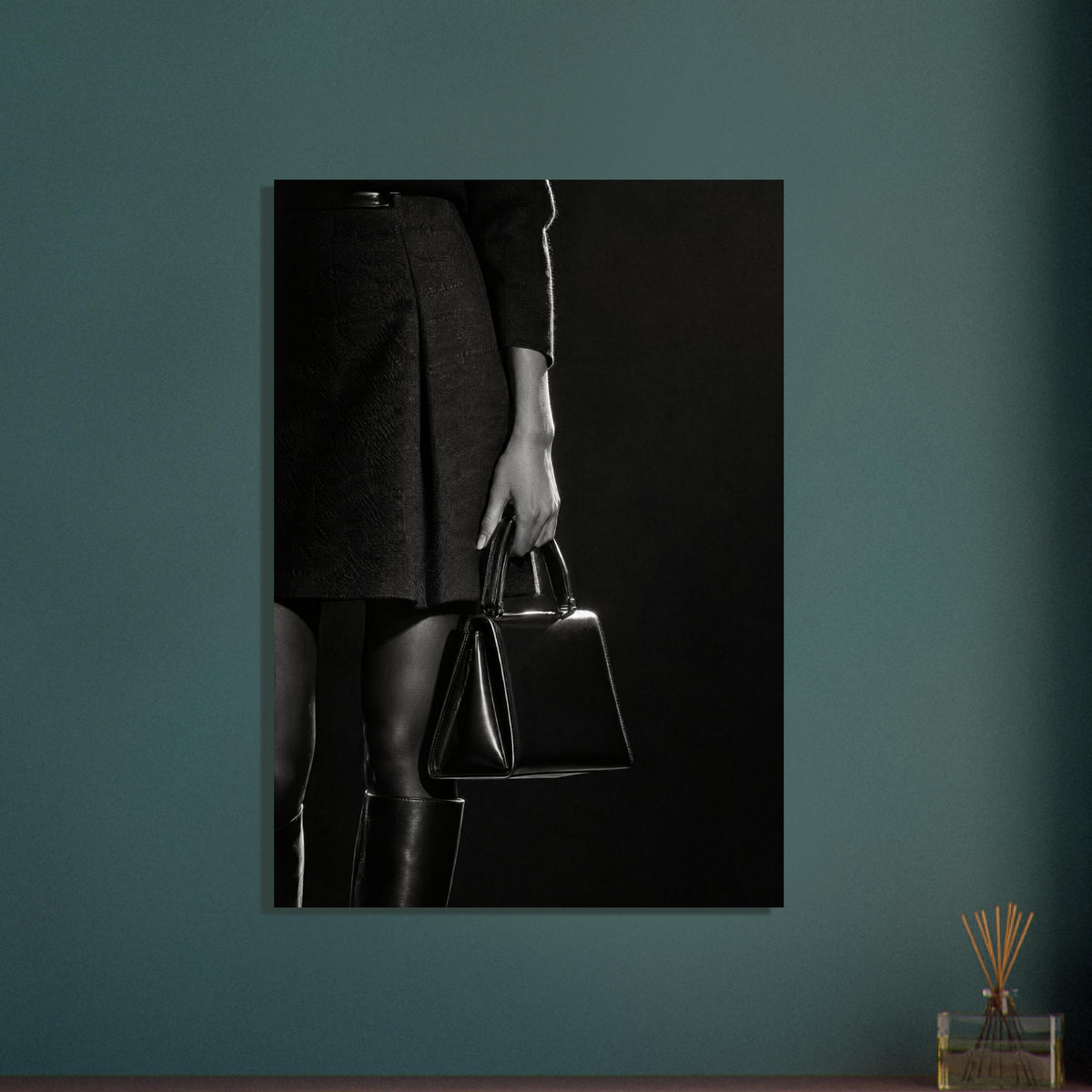 Cropped black and white photograph of a figure in a dark skirt and boots holding a glossy structured handbag.