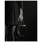 Cropped black and white photograph of a figure in a dark skirt and boots holding a glossy structured handbag.