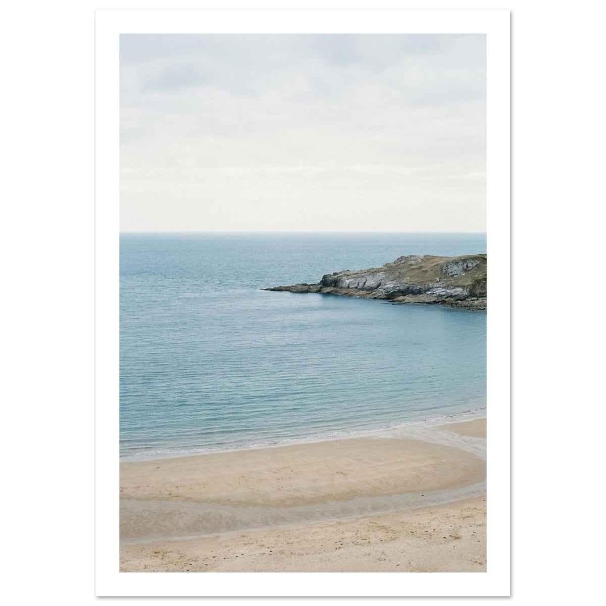 Sandy bay with calm blue water, a rocky headland and a curving tidal channel beneath a pale, overcast sky.