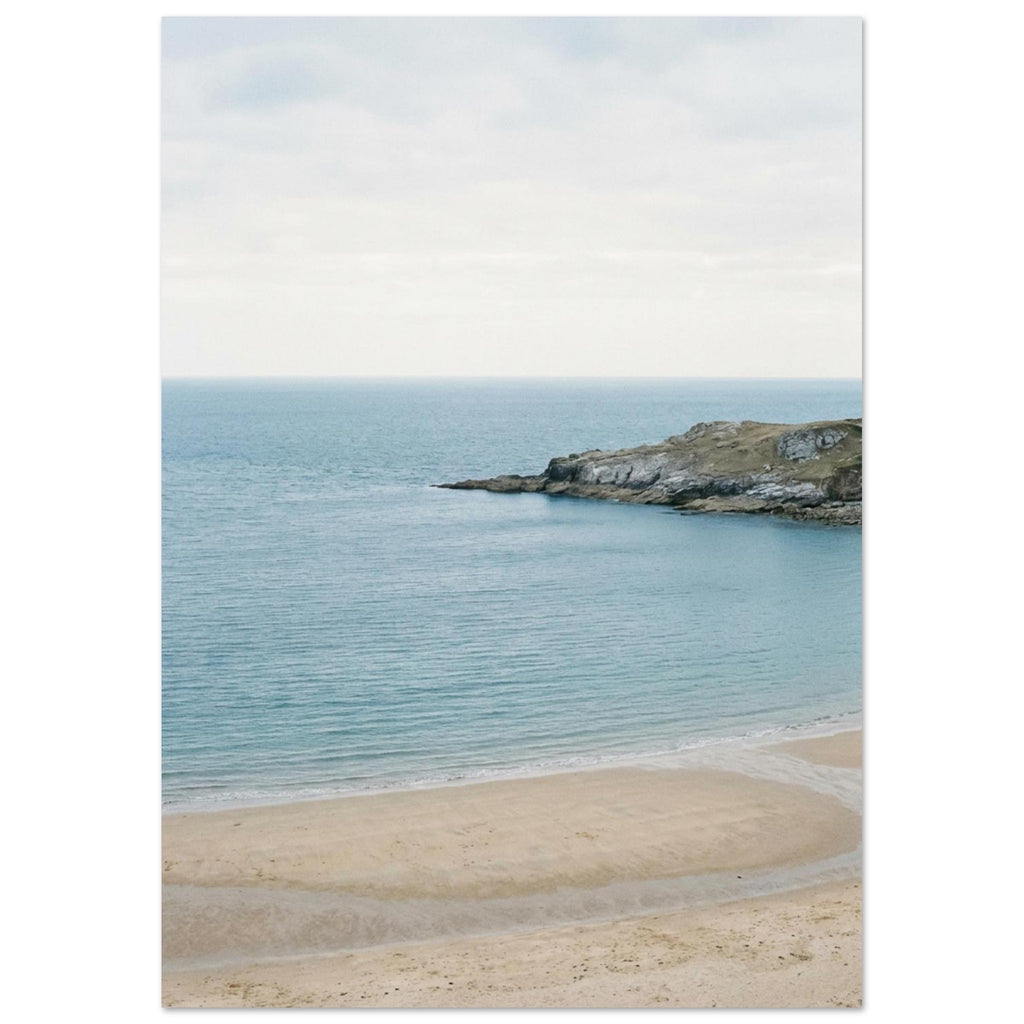 Sandy bay with calm blue water, a rocky headland and a curving tidal channel beneath a pale, overcast sky.