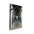 Timber pier viewed from beneath, with crossed wooden beams receding over gentle sea waves and wet sand under a grey sky.