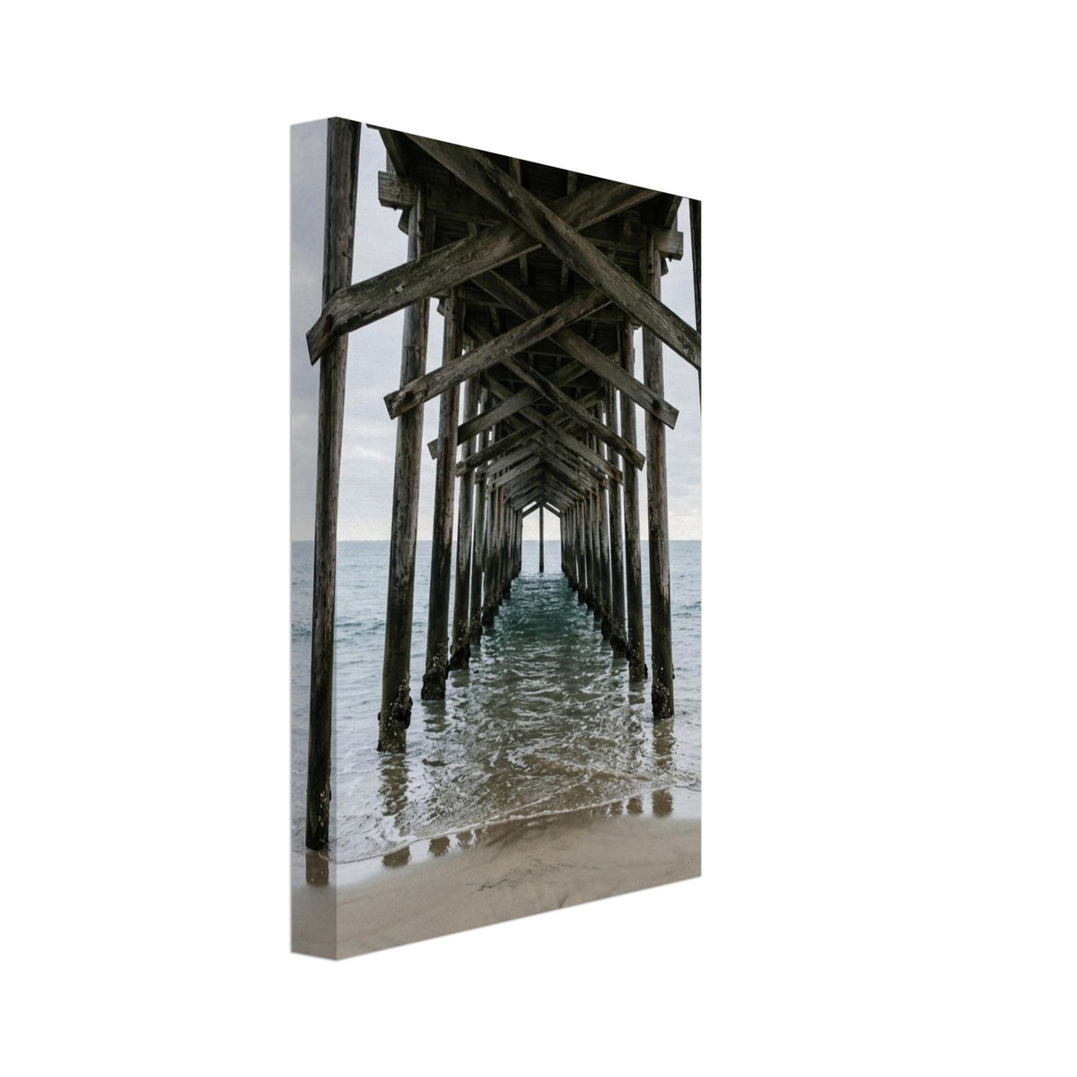 Timber pier viewed from beneath, with crossed wooden beams receding over gentle sea waves and wet sand under a grey sky.