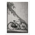 Bicycle leaning against a textured wall with long leafy shadows above and stone paving below, in black and white.