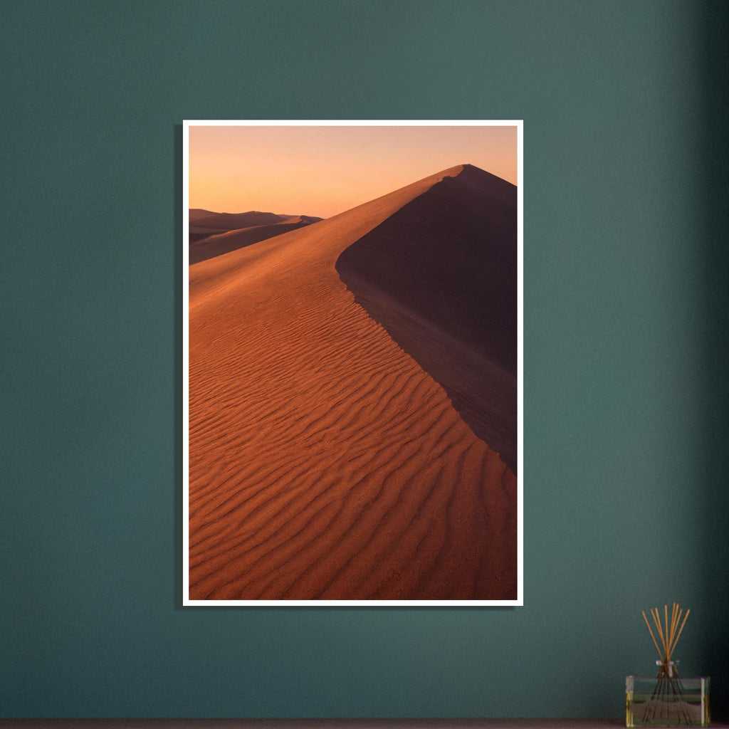 Desert sand dune with rippled orange slopes, a sweeping ridge and deep brown shadow beneath a peach sky.