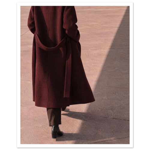 Figure seen from behind in a long burgundy coat, brown trousers and black boots on pink pavement with diagonal shadows.
