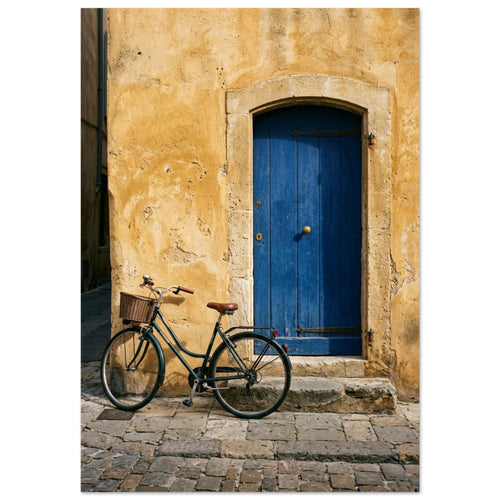 Green bicycle with a woven basket beside a blue wooden door in a golden plaster wall, above worn stone paving.