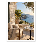 Sunlit stone terrace with a cushioned chair, round table and potted lemon tree overlooking blue sea and cliffs.