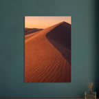 Desert sand dune with rippled orange slopes, a sweeping ridge and deep brown shadow beneath a peach sky.