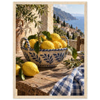 Lemons in a blue-and-white bowl on a wooden table, with olive branches, checked cloth and a coastal village beyond.