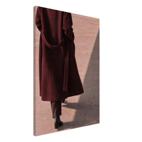 Figure seen from behind in a long burgundy coat, brown trousers and black boots on pink pavement with diagonal shadows.