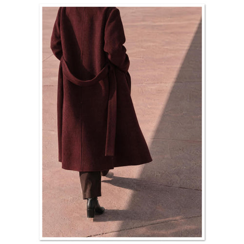 Figure seen from behind in a long burgundy coat, brown trousers and black boots on pink pavement with diagonal shadows.