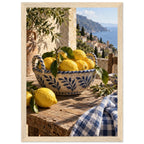 Lemons in a blue-and-white bowl on a wooden table, with olive branches, checked cloth and a coastal village beyond.