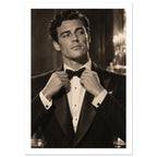 Man in a dark dinner jacket adjusting his bow tie, looking sideways, with blurred lights behind him in warm sepia tones.