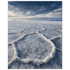 Salt flats with raised polygon-shaped ridges stretching towards distant mountains beneath a blue, cloud-streaked sky.