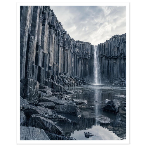Waterfall cascading between tall basalt columns into a reflective pool, with dark foreground rocks and a cloudy sky.