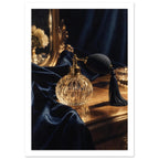 Faceted glass perfume bottle with black bulb and tassel on a golden tabletop beside navy drapery and an ornate mirror.