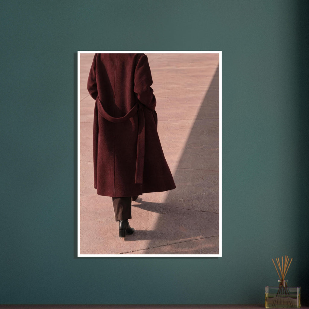 Figure seen from behind in a long burgundy coat, brown trousers and black boots on pink pavement with diagonal shadows.