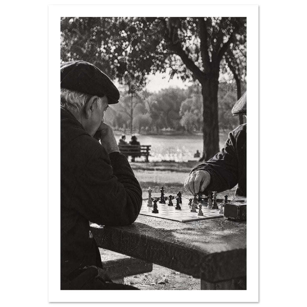 Two men in flat caps playing chess at a stone table beside water and trees, photographed in black and white.