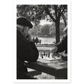 Two men in flat caps playing chess at a stone table beside water and trees, photographed in black and white.