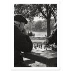 Two men in flat caps playing chess at a stone table beside water and trees, photographed in black and white.