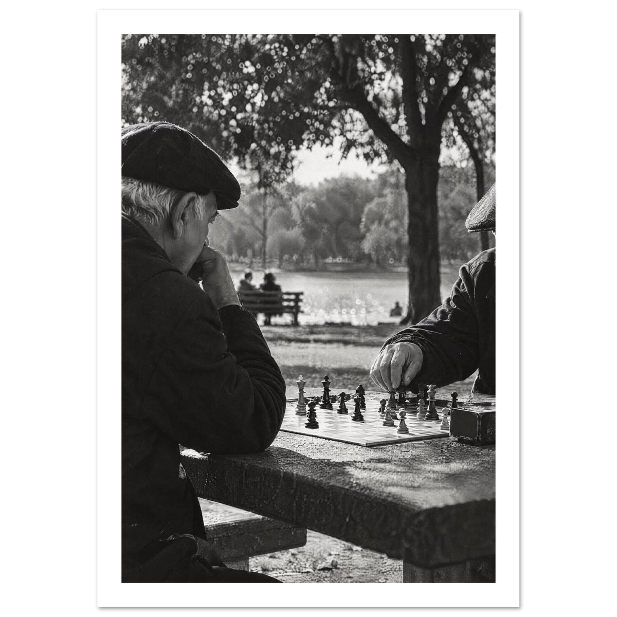 Two men in flat caps playing chess at a stone table beside water and trees, photographed in black and white.