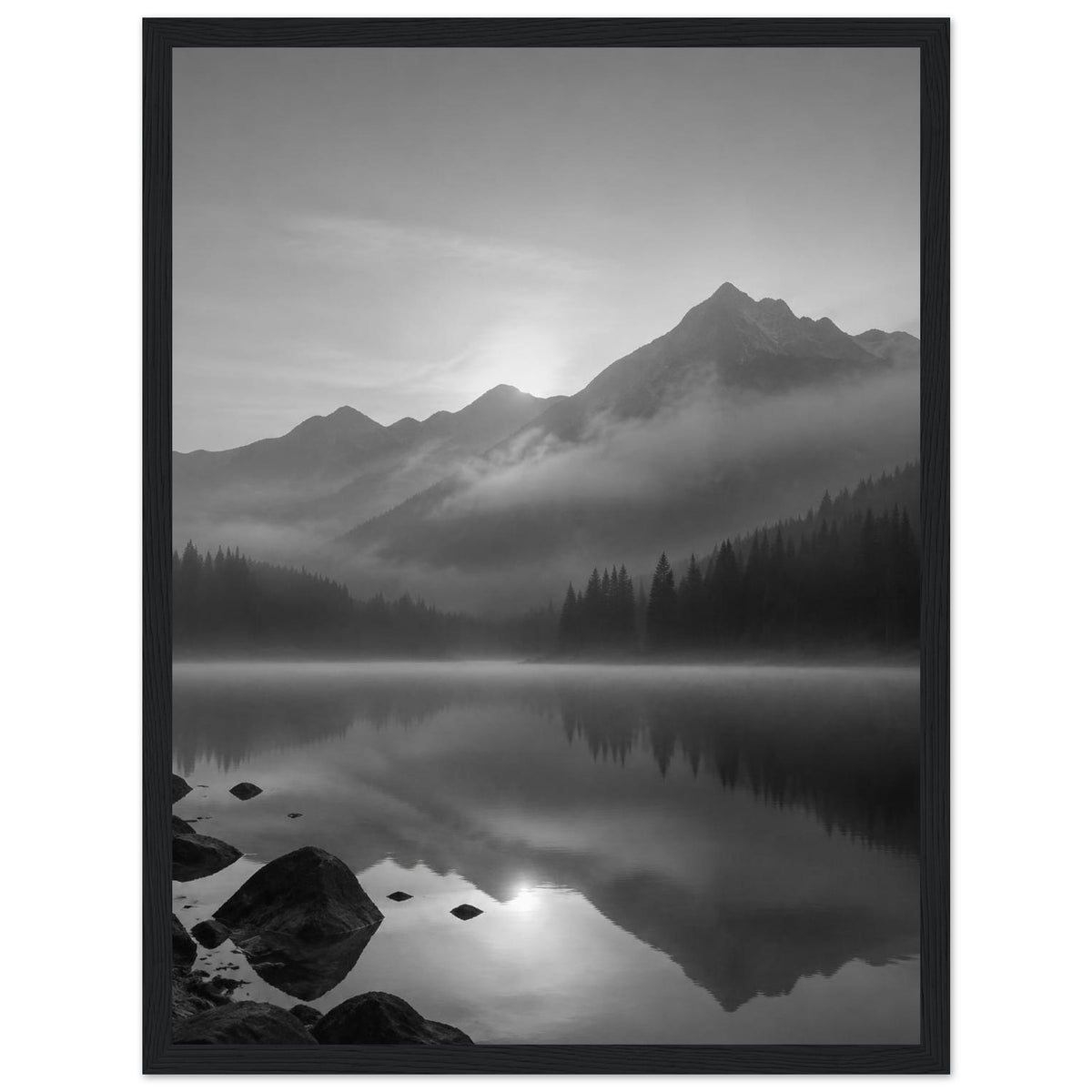 Mountain peaks and conifer forests reflected in a misty lake, with rocks in the foreground, in black and white.