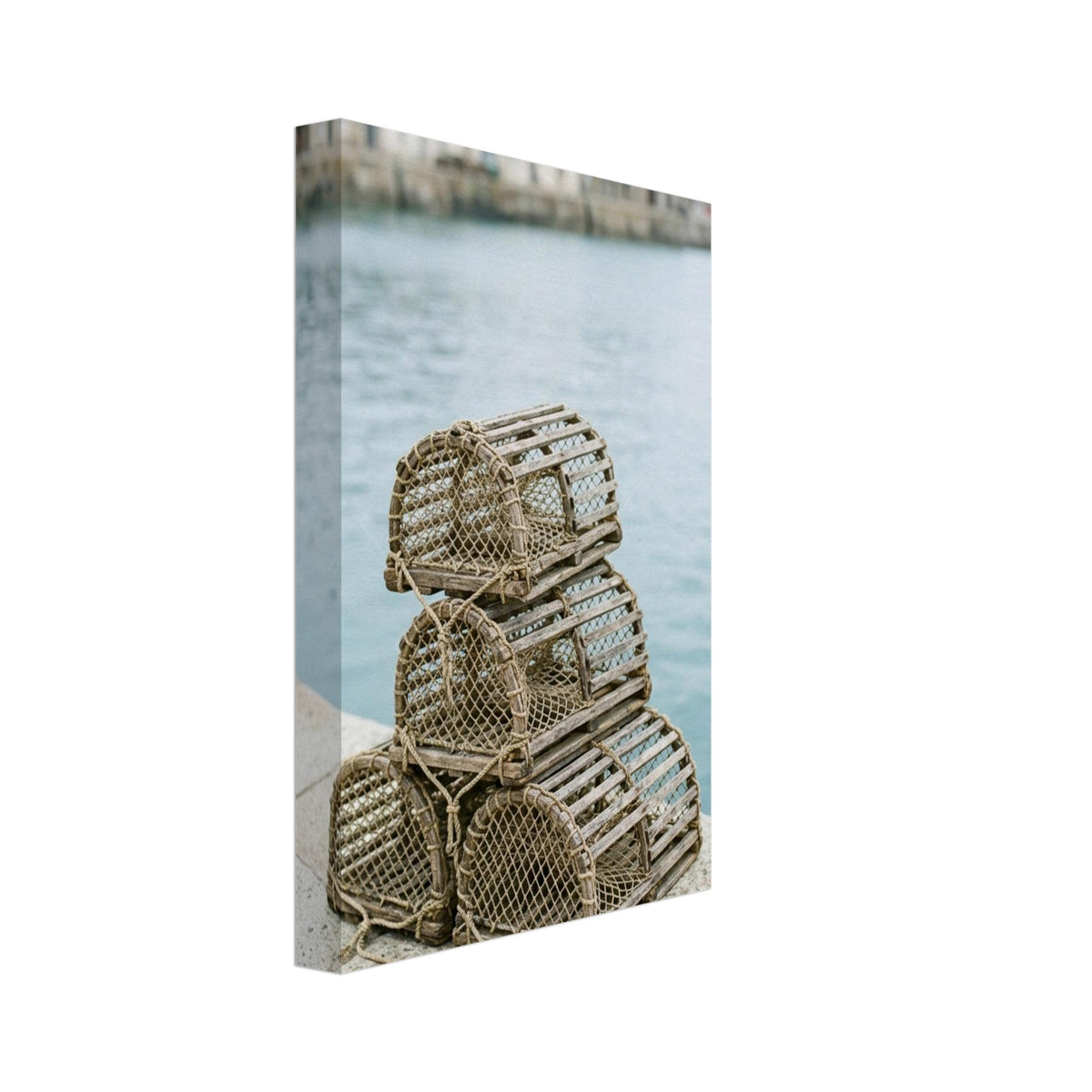 Four wooden lobster pots stacked on a stone quay beside pale blue harbour water, with blurred waterfront buildings beyond.