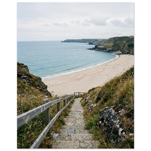 Stone steps with a wooden handrail descend through coastal grasses to a sandy beach, blue sea and rocky headlands.