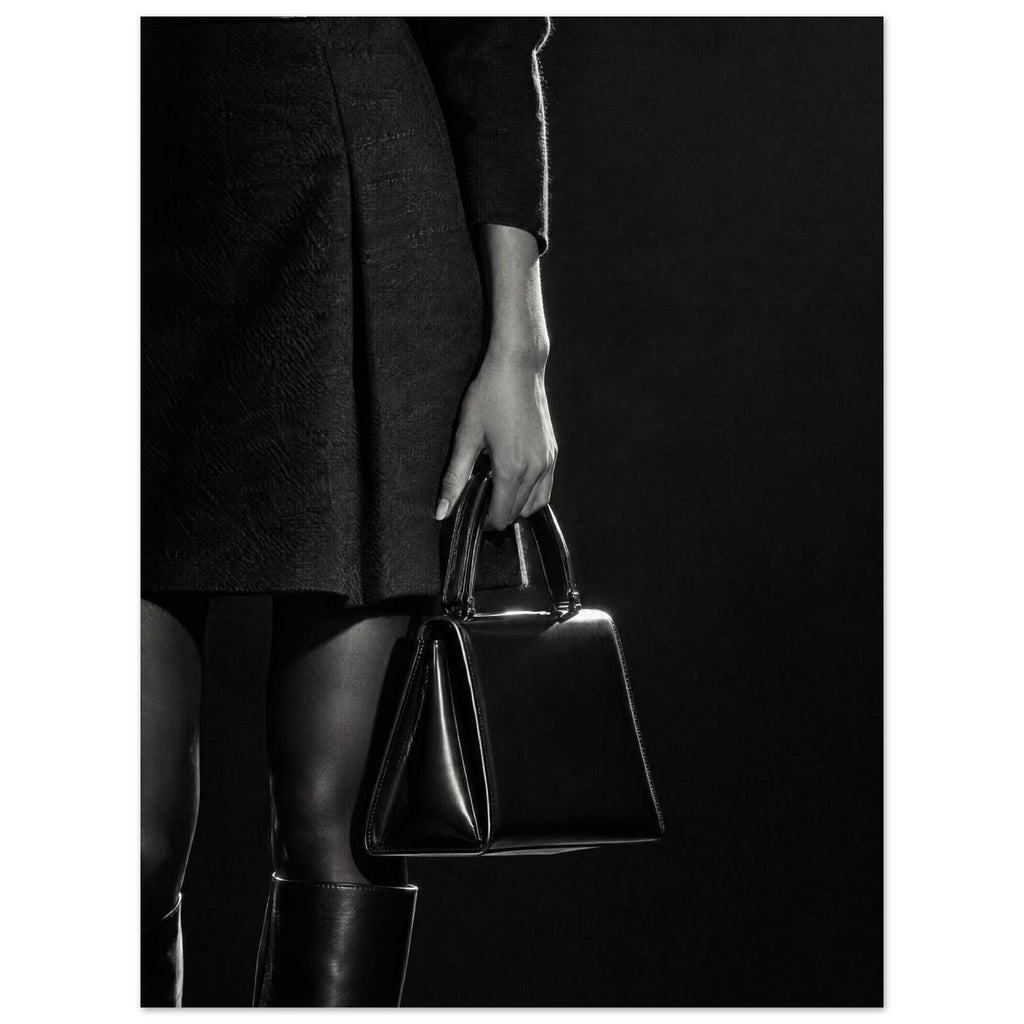 Cropped black and white photograph of a figure in a dark skirt and boots holding a glossy structured handbag.