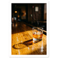 Tumbler of amber drink with ice on a sunlit wooden bar, casting a long shadow against a dark, blurred interior.