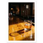 Tumbler of amber drink with ice on a sunlit wooden bar, casting a long shadow against a dark, blurred interior.