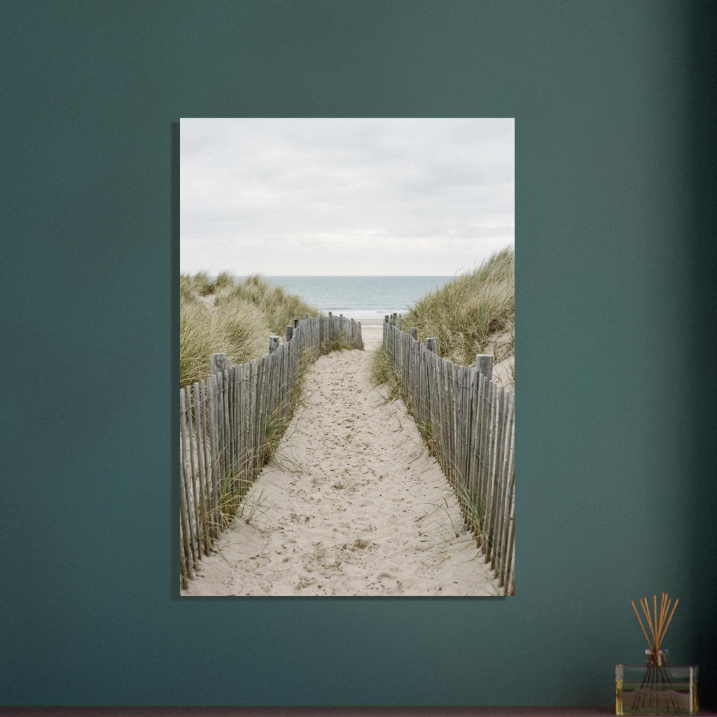 Sandy path with footprints between wooden fences and grassy dunes, leading towards a pale blue sea beneath a cloudy sky.