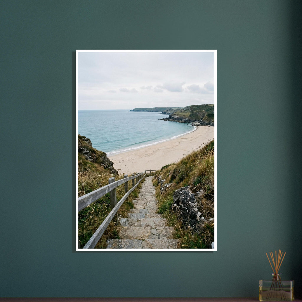 Stone steps with a wooden handrail descend through coastal grasses to a sandy beach, blue sea and rocky headlands.