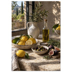 Lemons, olives, figs and a bottle of oil on a stone table beside a ceramic jug of leafy branches and a sunlit window.